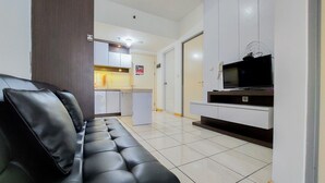 A living room with a black leather sofa, a flat-screen TV on a stand, and a kitchen area in the background. - Warm And Nice 2Br Apartment At M-Town Residence (Gading Serpong)