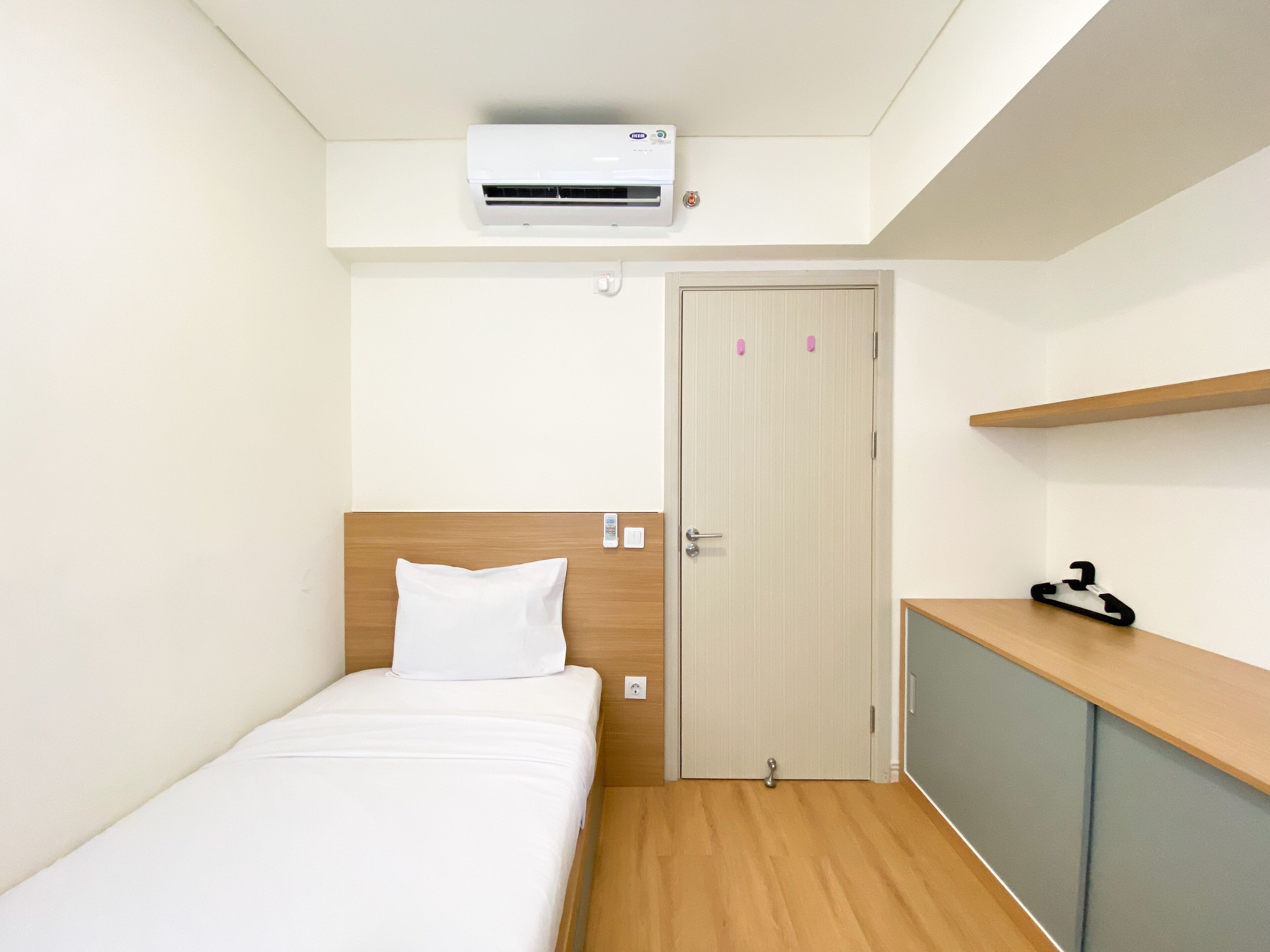 A small, neatly arranged bedroom with a single bed, a wooden headboard, a wardrobe, a door with a handle, and an air conditioning unit mounted on the wall.