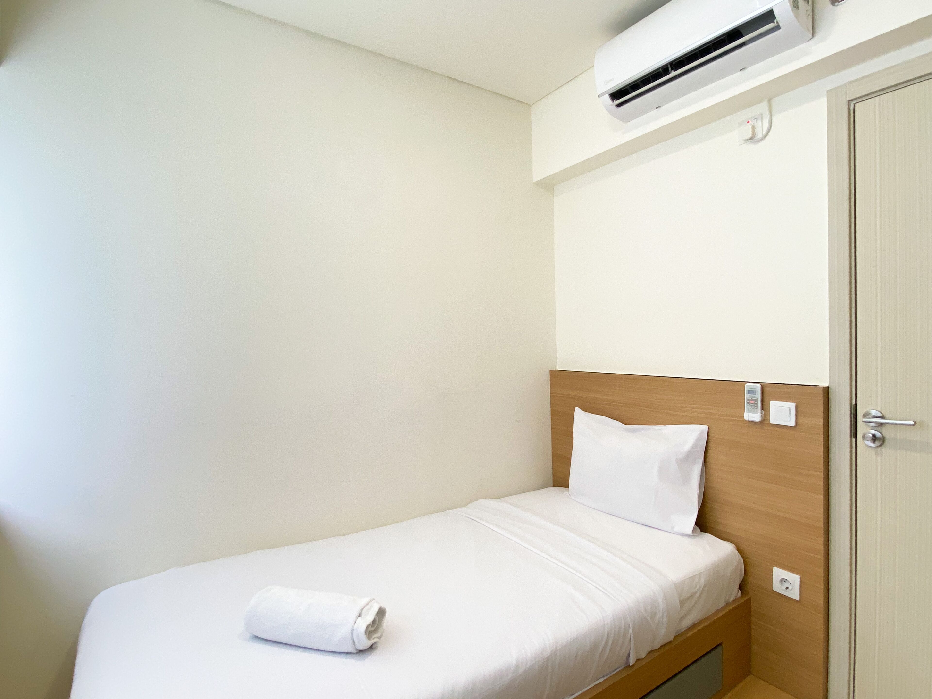 A single bed with a wooden headboard, white bedding, and a rolled towel. An air conditioning unit is mounted on the wall above the bed.