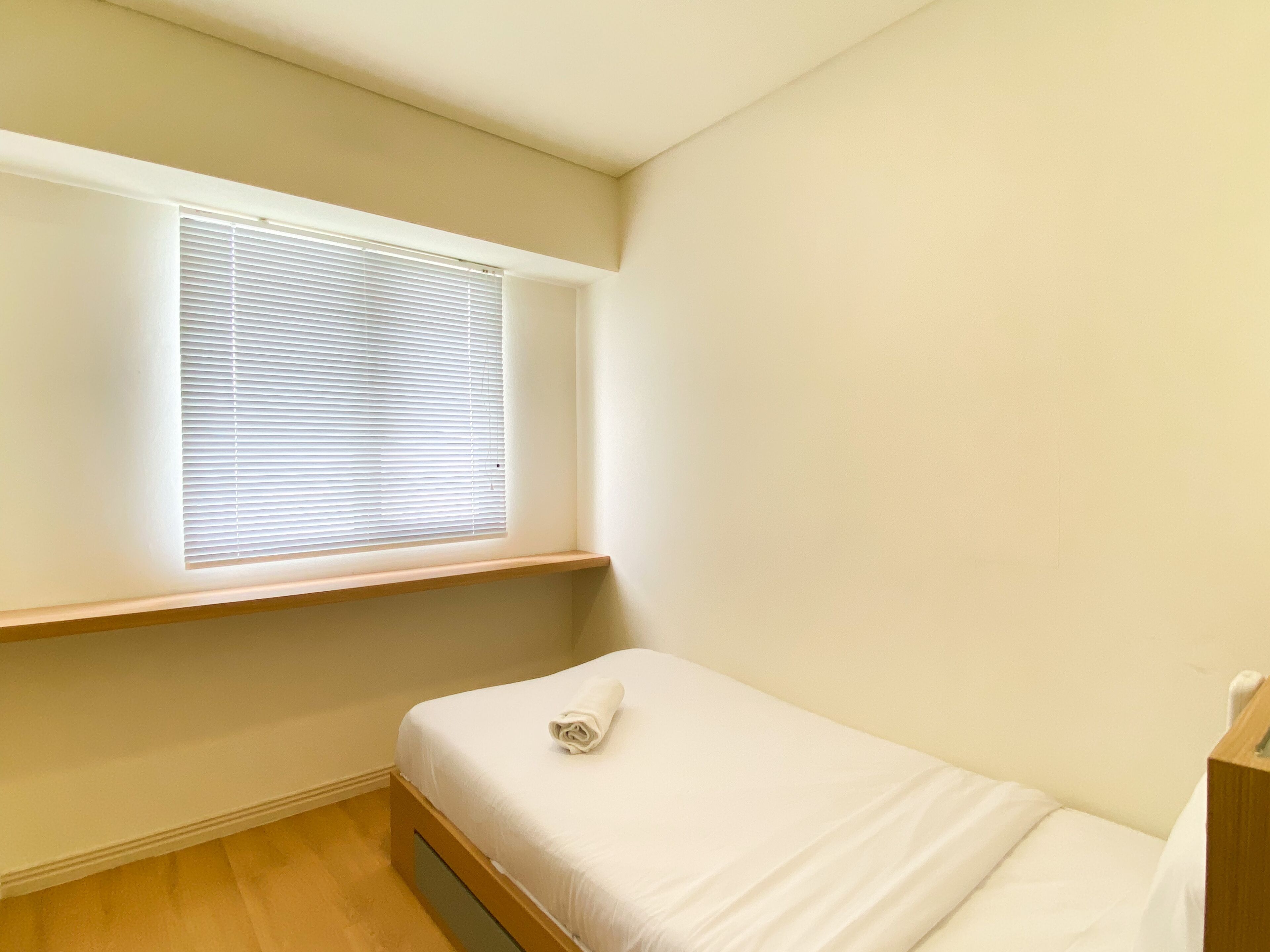 A small, neatly arranged room with a single bed, a window with blinds, and a wooden shelf.