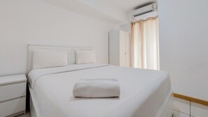 Una cama bien hecha con edredón blanco y una toalla enrollada, una mesita de noche y una ventana con cortinas.