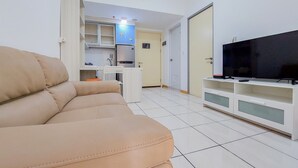 Una sala de estar con un sofá color beige, un televisor sobre un mueble blanco, y una cocina al fondo.