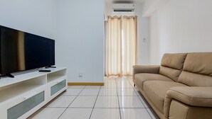 Una sala de estar con una televisión de pantalla plana sobre un mueble blanco, un sofá color beige y una ventana con cortinas.