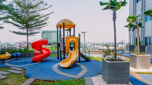 Ein Spielplatz mit zwei Rutschen und einer Kletterkonstruktion, umgeben von Grünflächen und Palmen.
