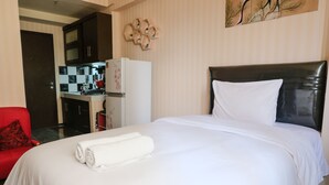 A hotel room with a bed, a red chair, a kitchen area, and a painting on the wall. - Homey And Cozy Studio Room At Suites @Metro Apartment (Bandung)