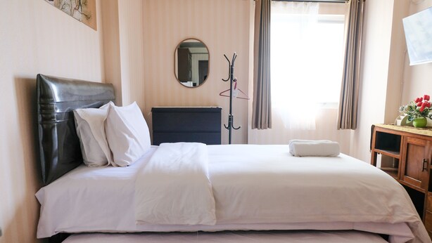 A hotel room with a bed, a wooden dresser, a coat rack, and a mirror on the wall. - Homey And Cozy Studio Room At Suites @Metro Apartment (Bandung)