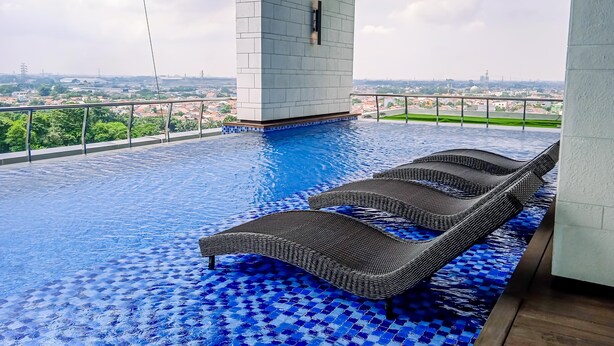 An outdoor infinity pool with two wicker lounge chairs, a tiled floor, and a view of the cityscape. - Spacious And Elegant 2Br At 35Th Floor Hillcrest House Apartment (Curug)