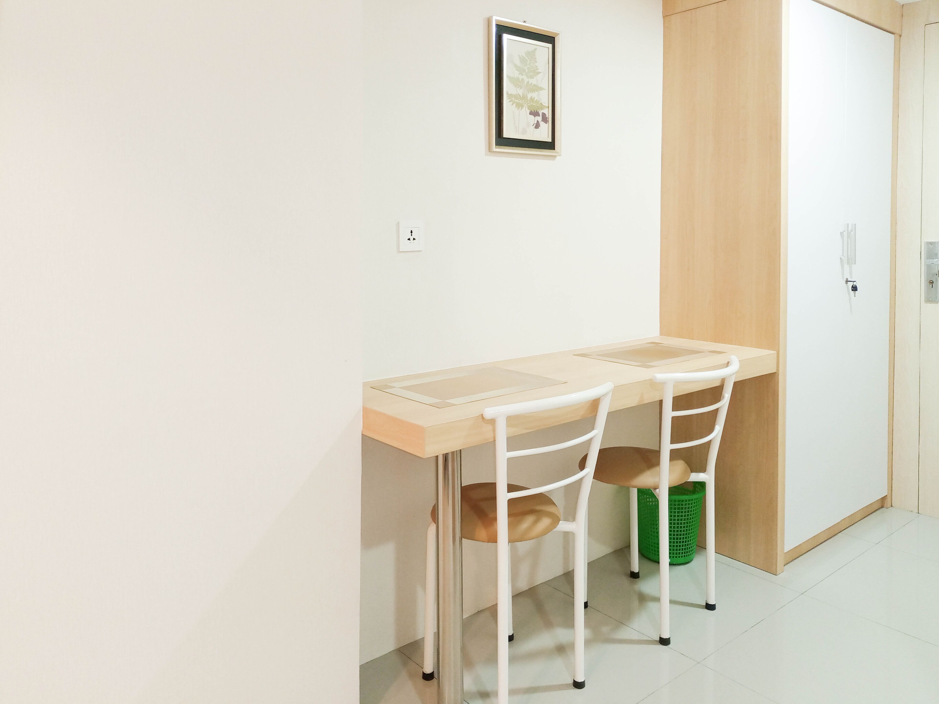 A small, clean room with a wooden desk and two chairs, a green laundry basket, and a door with a handle.