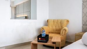 A hotel room with a wooden armchair, a small wooden table with a water bottle and a box labeled "Travelio," and a mirror on the wall.