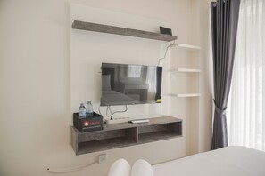 A hotel room with a flat-screen TV mounted on the wall, a small table with a remote control, a water bottle, and a tray of snacks. - Good Deal And Homey Studio At 11St Floor Citra Living Apartment (Jakarta)