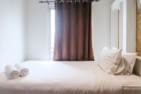Un lit bien fait, avec du linge de lit blanc et des oreillers, une fenêtre avec des rideaux bruns et un mur blanc.