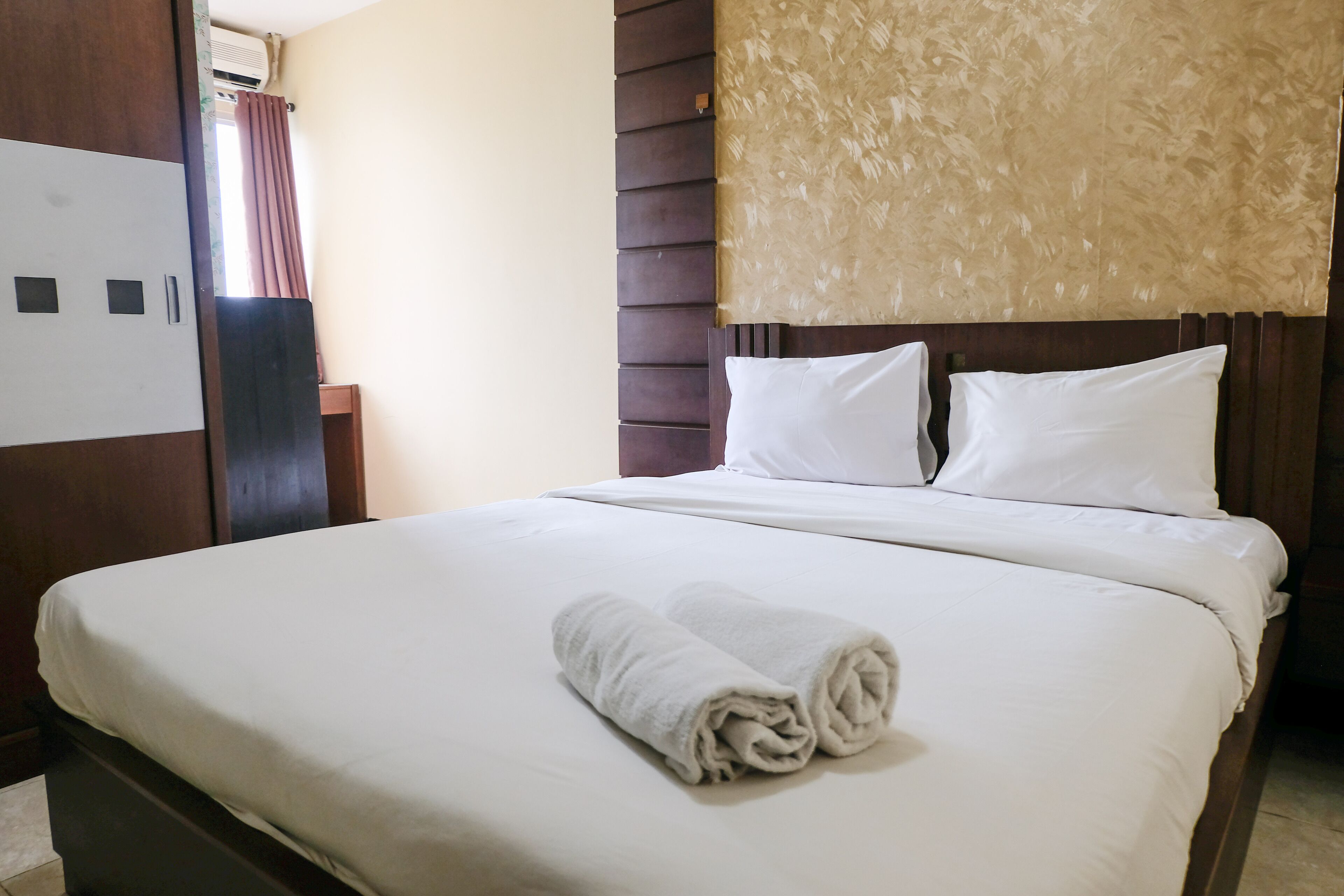 A neatly made bed with white linens and a rolled towel, a wooden headboard, and a beige textured wall.