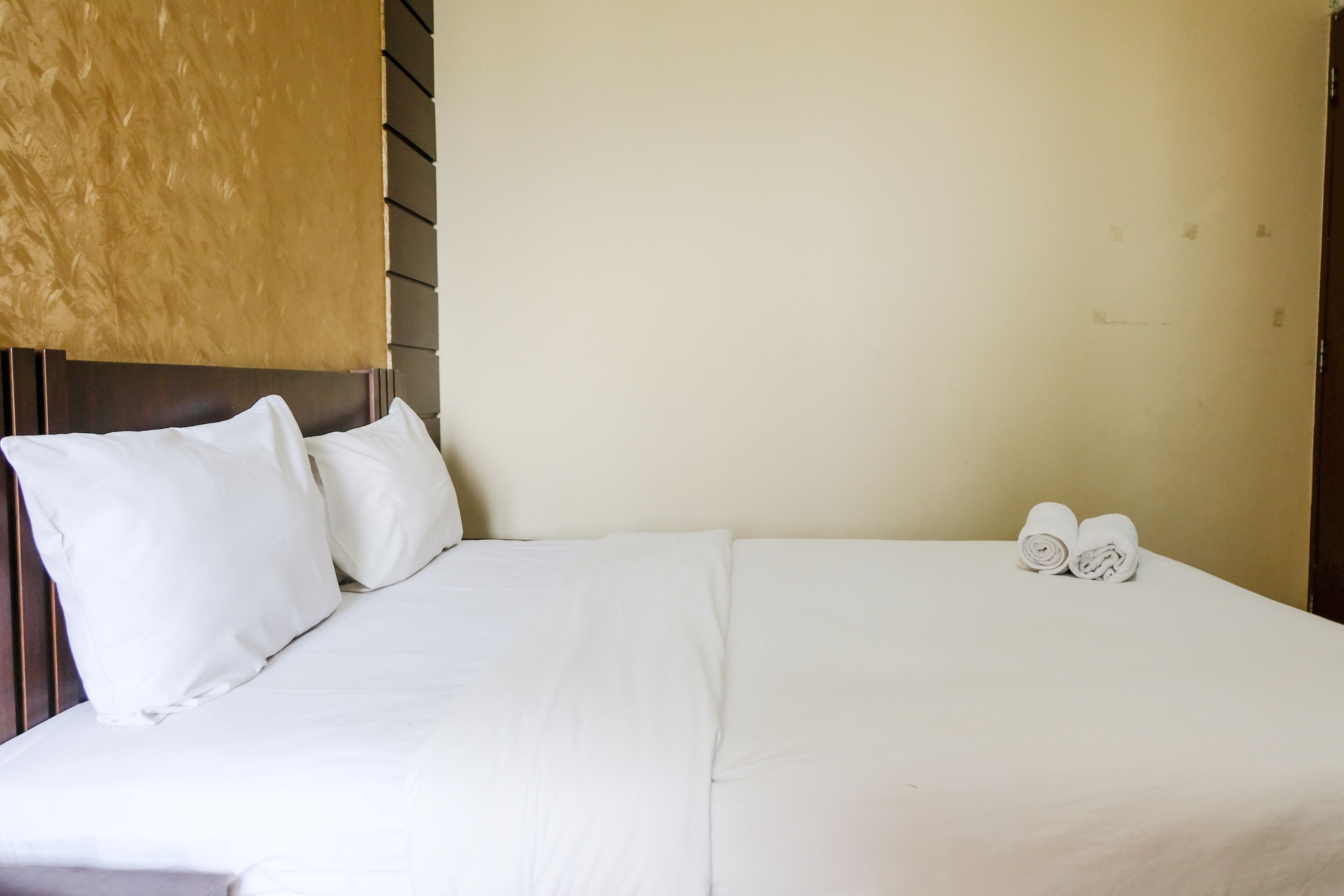 A neatly made bed with white linens and pillows, a headboard with a textured panel, and two rolled towels on the bed.