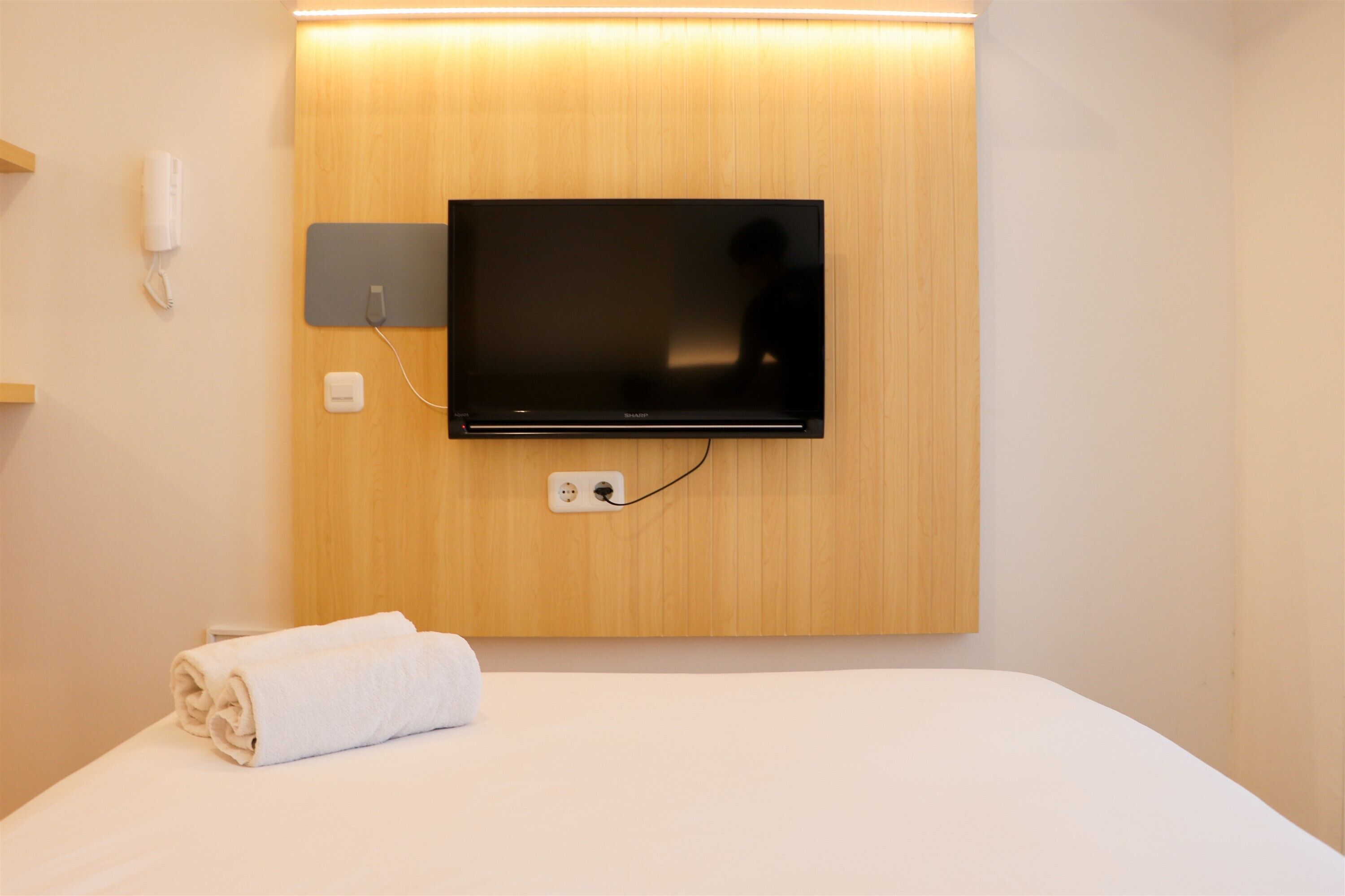 A hotel room with a flat-screen TV mounted on the wall, a rolled white towel on the bed, and a power outlet on the wall.