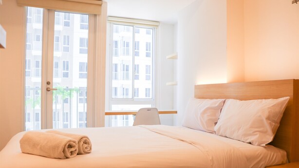 A hotel room with a bed, a nightstand, a chair, and a window with a view of buildings. - Modern Studio Room At Tokyo Riverside Pik 2 Apartment (Teluknaga)