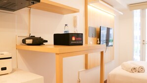 A hotel room with a wooden table, a black travel bag, a water bottle, a white appliance, a flat-screen TV, and a bed with a rolled towel. - Modern Studio Room At Tokyo Riverside Pik 2 Apartment (Teluknaga)