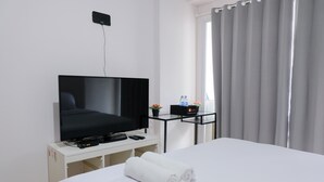 A hotel room with a bed, a TV on a white stand, a small table with a remote control, a water bottle, and a potted plant. - Homey Studio Room At 7Th Floor Gateway Pasteur Apartment (Cimahi)