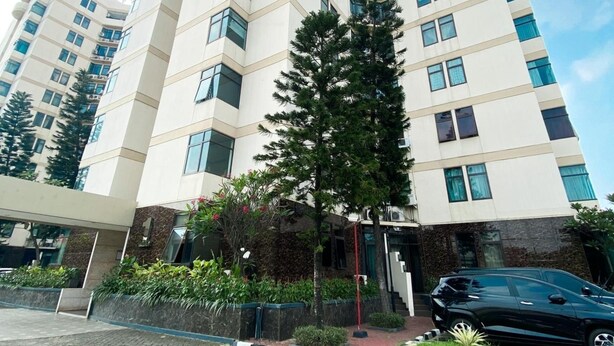 Un moderno edificio de gran altura con fachada blanca, varios balcones y un área de entrada ajardinada con un gran árbol y arbustos.
