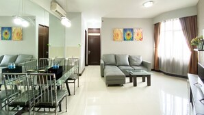 Una sala moderna con una mesa de vidrio, un sofá gris y una mesa de centro. Hay tres cuadros en la pared, un ventilador de techo y una puerta que da a otra habitación.