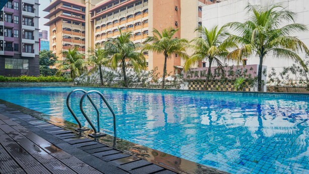 A swimming pool with a wooden deck, surrounded by palm trees and modern buildings. - Modern And Cozy Stay 1Br At Tamansari Semanggi Apartment (Jakarta)