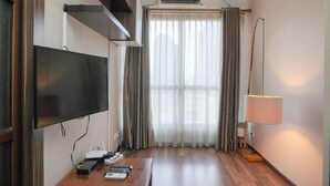 A room with a flat-screen TV, an air conditioner, a standing lamp, a wooden desk with remote controls, and a window with curtains. - Modern And Cozy Stay 1Br At Tamansari Semanggi Apartment (Jakarta)