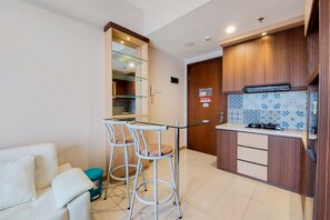 A compact kitchen with a glass-top table, two bar stools, a white sofa, and a wooden cabinet with a patterned backsplash. - Homey And Simply 1Br M-Town Signature Apartment (Gading Serpong)