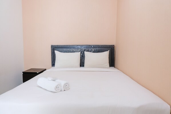 A neatly made bed with white linens and two pillows, a small bedside table, and a wall with a light peach color.