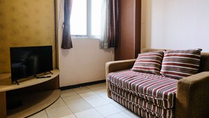 A living room with a flat-screen TV on a wooden stand, a striped sofa, and a window with curtains. - Homey 2Br At Suites @Metro Apartment (Bandung)