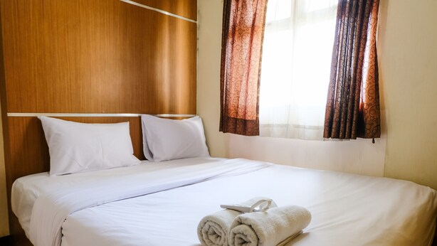 A neatly made bed with white linens and two rolled towels, a wooden headboard, and a window with patterned curtains. - Homey 2Br At Suites @Metro Apartment (Bandung)