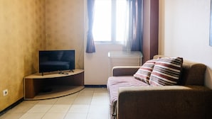 A small, cozy room with a sofa, a television on a stand, a radiator, and a window with curtains. - Homey 2Br At Suites @Metro Apartment (Bandung)