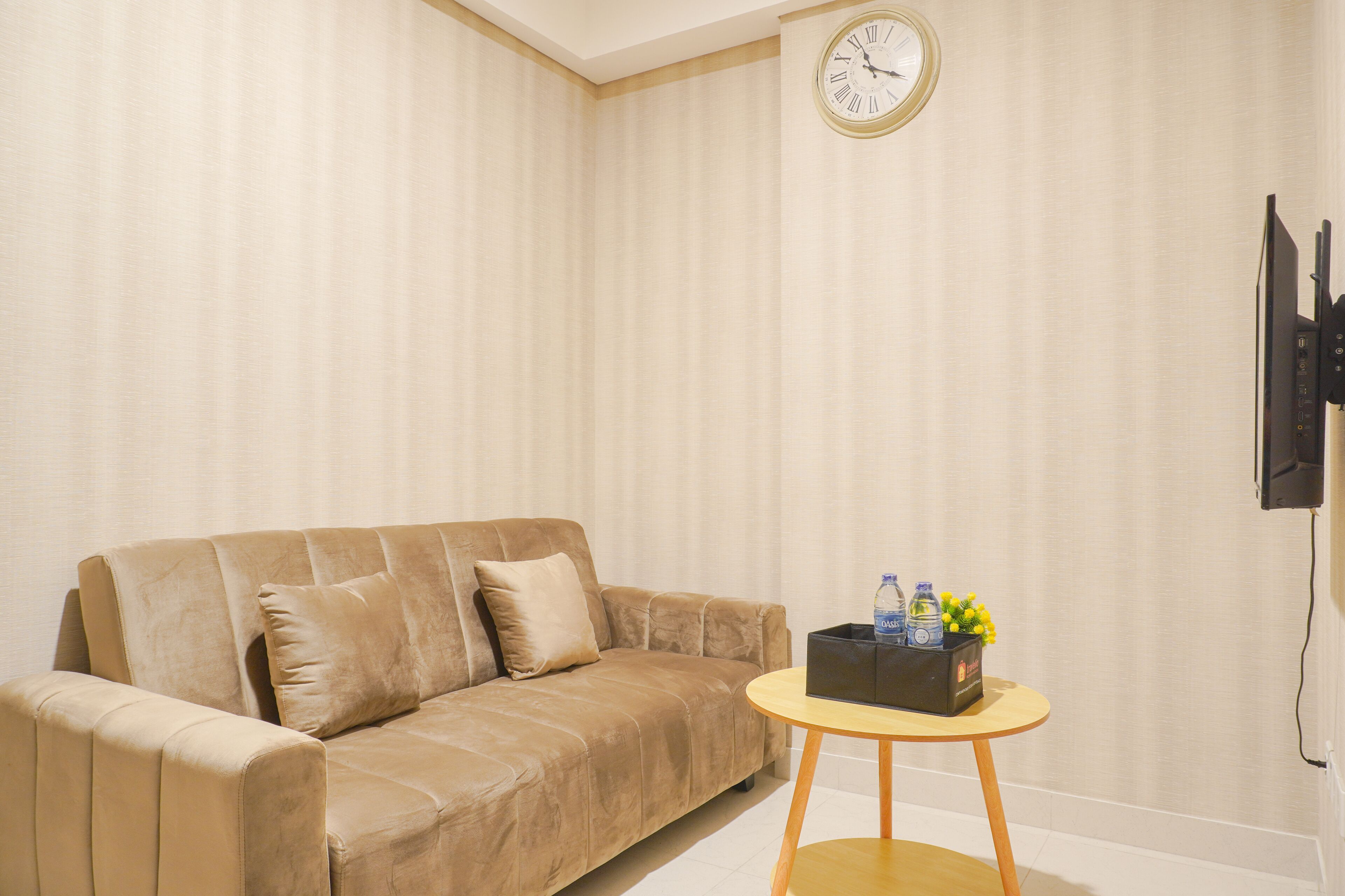 Ein beiges Sofa mit zwei Kissen, ein runder Holztisch mit einer schwarzen Box, die Wasserflaschen und Blumen enthält, und eine an der Wand befestigte Uhr.