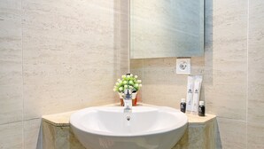 A bathroom with a large white sink, a mirror above it, and a vase with white flowers. There are toiletries on the counter. - Modern Look 1Br With Golf Island And Sea View Gold Coast Apartment (Jakarta)