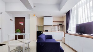 A compact living space with a glass-top dining table, a sofa, a kitchen area, and a television. - Restful And Modern 2Br Majestic Point Serpong Apartment (Gading Serpong)