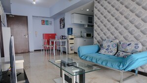 A modern living room with a glass coffee table, a blue sofa with patterned cushions, a red table, and a kitchen area in the background. - Homey And Cozy 1Br Apartment At Braga City Walk (Bandung)