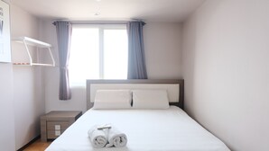 A neatly made bed with white linens and rolled towels, a bedside table with a lamp, and a window with curtains. - Homey And Cozy 1Br Apartment At Braga City Walk (Bandung)