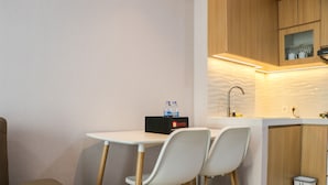 A compact kitchen with a white countertop, two white chairs, a small trash bin, and wooden cabinets. - Brand New 2BR at Daan Mogot City Apartment By Travelio (Jakarta)