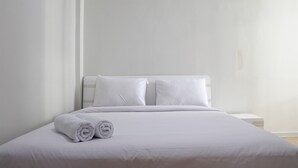 A neatly made bed with white linens and pillows, a small bedside table, and a plain wall. - Brand New 2BR at Daan Mogot City Apartment By Travelio (Jakarta)