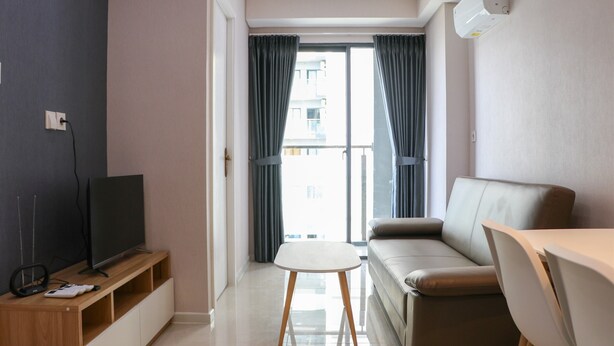 A modern living room with a flat-screen TV on a wooden stand, a beige sofa, a small white table, and a sliding glass door leading to a balcony. - Brand New 2BR at Daan Mogot City Apartment By Travelio (Jakarta)