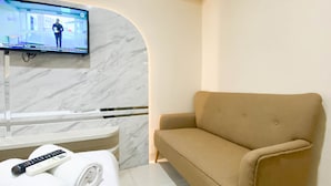 Une chambre d’hôtel avec un canapé beige, un téléviseur fixé sur un mur de marbre, une table de massage et une télécommande posée sur une serviette.