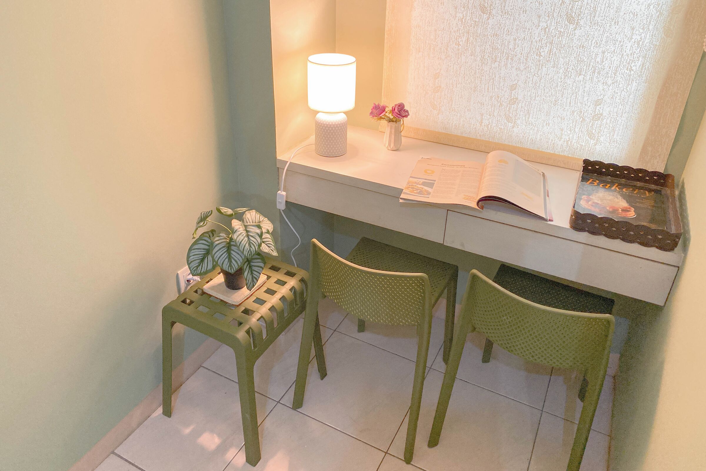 Un petit espace de travail comprenant un bureau, deux chaises, une lampe, une plante et une fenêtre ornée de rideaux en dentelle.