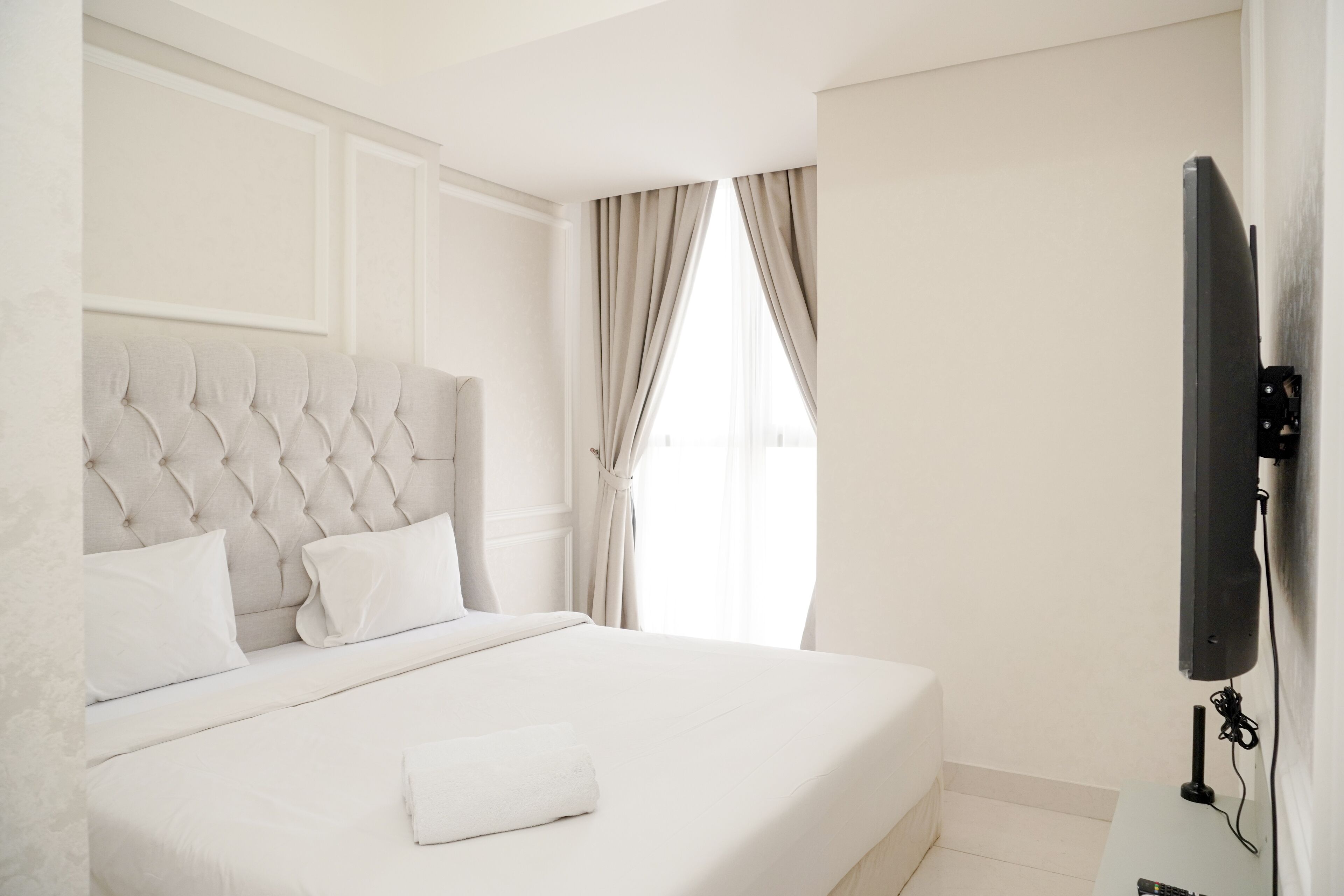 A hotel room with a tufted headboard, a flat-screen TV mounted on the wall, and a large window with curtains.
