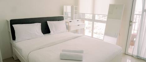 A neatly made bed with white linens and a folded towel, a bedside table with a mirror, and a view of the outdoors through a large window.
