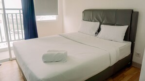 A neatly made bed with white linens and a folded towel, a grey upholstered headboard, and a window with a view of buildings. - Minimalist And Best Deal Studio At Serpong Garden Apartment (Cisauk)
