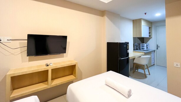 A hotel room with a bed, a TV mounted on the wall, a wooden shelf, a kitchenette, and a chair. - Simply Look Studio At 21St Floor De Prima Apartment (Medan)