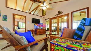 A spacious living room with wooden furniture, a ceiling fan, and a large window. - Entire Beach Home w/ Sunroom, Pool, Bikes, Paddleboards, BBQ & more (Hopkins)