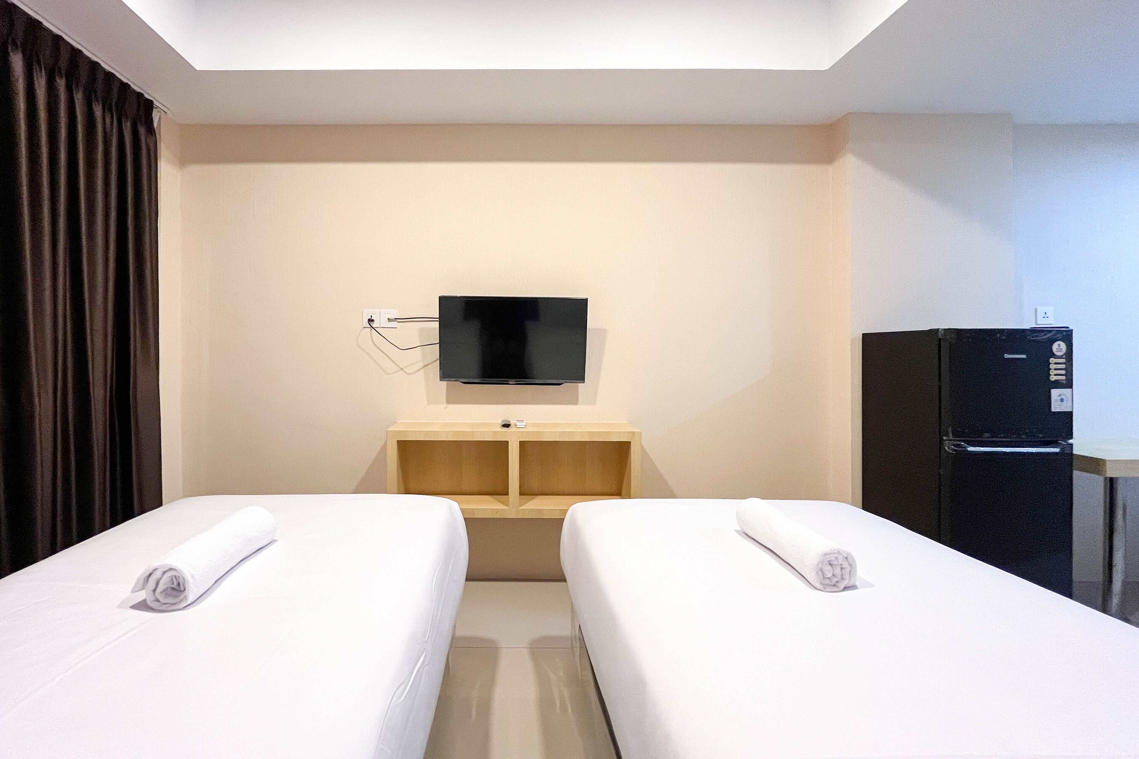 A hotel room with two beds, a TV mounted on the wall, a small wooden cabinet, and a refrigerator.