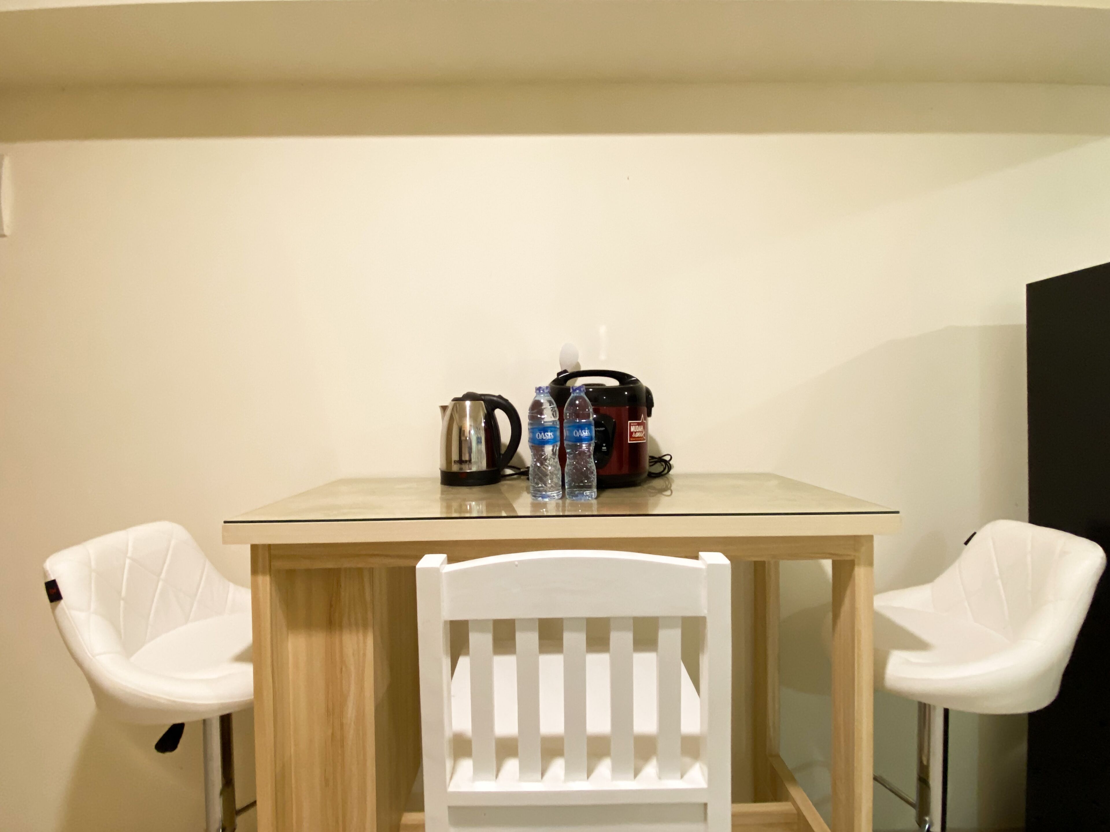 A small dining area with a glass-top table, two white chairs, a kettle, a water bottle, and a rice cooker.