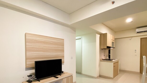 A modern living room with a flat-screen TV on a wooden console, a kitchen area with wooden cabinets, and a hallway leading to another room. - Comfortable And Nice 2Br Apartment At Meikarta (Cikarang)