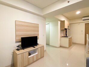 A modern living room with a flat-screen TV on a wooden console, a kitchen area with wooden cabinets, and a hallway leading to another room.