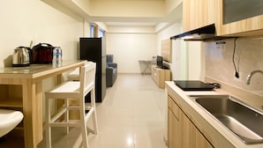 A compact kitchen with wooden cabinets, a stainless steel sink, a refrigerator, and a dining area with a chair and table. - Comfortable And Nice 2Br Apartment At Meikarta (Cikarang)